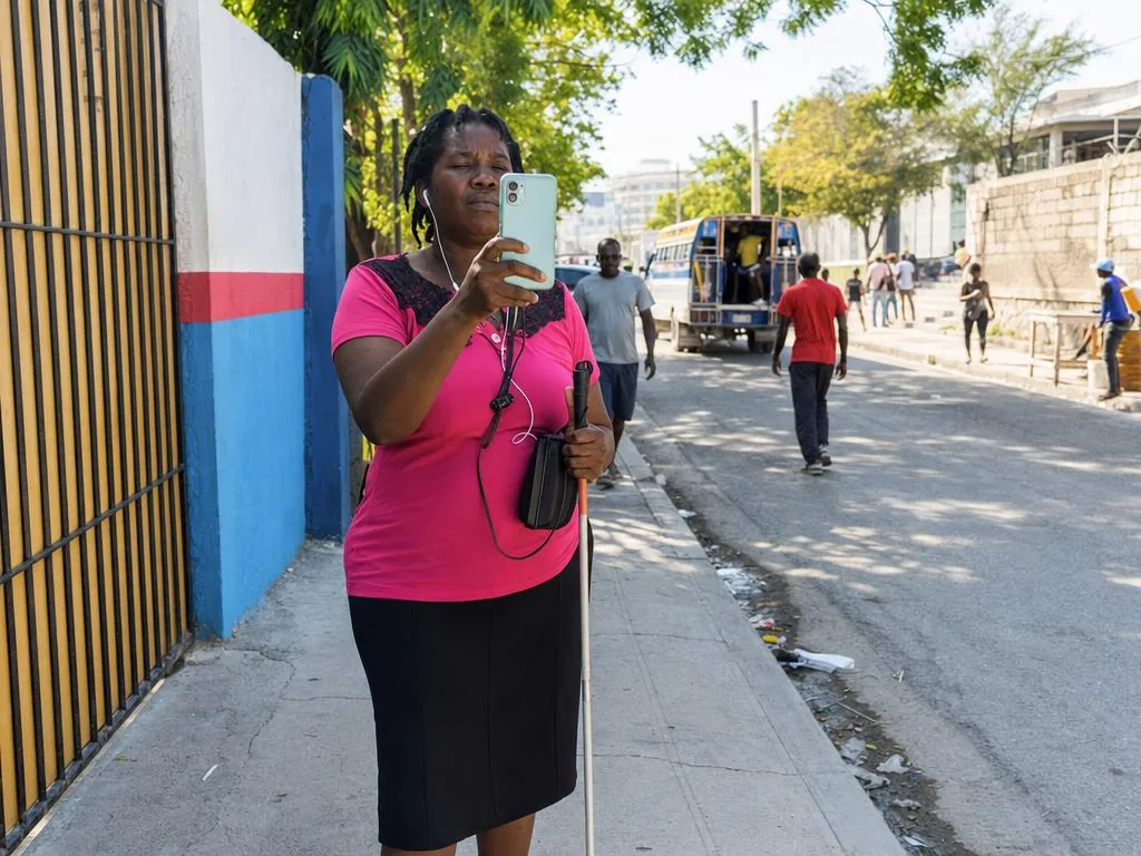 Une femme avec une canne blanche tient son téléphone droit devant sa poitrine, la caméra tournée vers la rue.